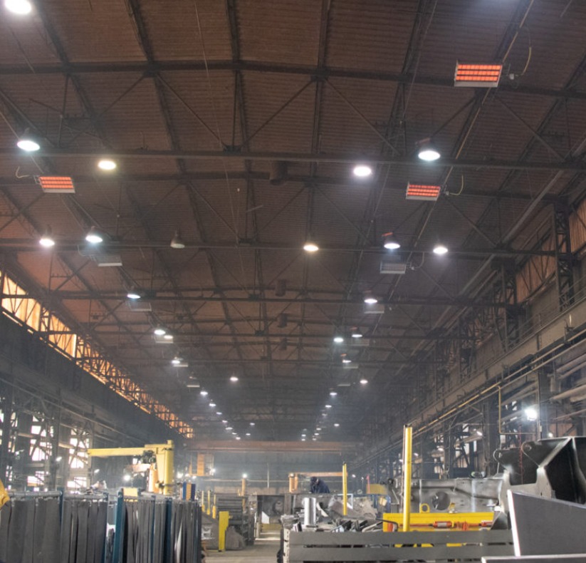 Wnętrze hali przemysłowej z widocznymi elementami instalacji grzewczej pod sufitem, oświetleniem i ciężkimi maszynami.