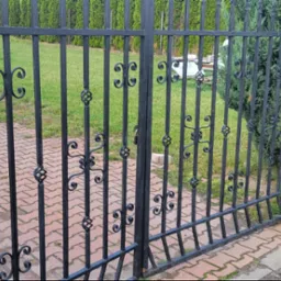 Czarna metalowa brama z ozdobnymi elementami w kształcie spirali i wolut, przylegająca do ceglanego słupka i zielonego żywopłotu, z widoczną kostką brukową i trawnikiem w tle.