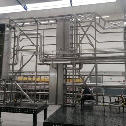 MICHAŁ GĄSIOROWSKI „SDC” - Specjalista dla Ciebie - Konstrukcja ze stali nierdzewnej w hali produkcyjnej, widoczne rury, spawane elementy, system zaworów, stoły spawalnicze z otworami, w tle regały magazynowe i pracownik.