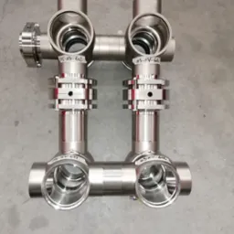 Precyzyjnie wykonane, symetryczne złącze rurowe ze stali nierdzewnej z widocznymi oznaczeniami na elementach, leżące na szarym, betonowym podłożu.