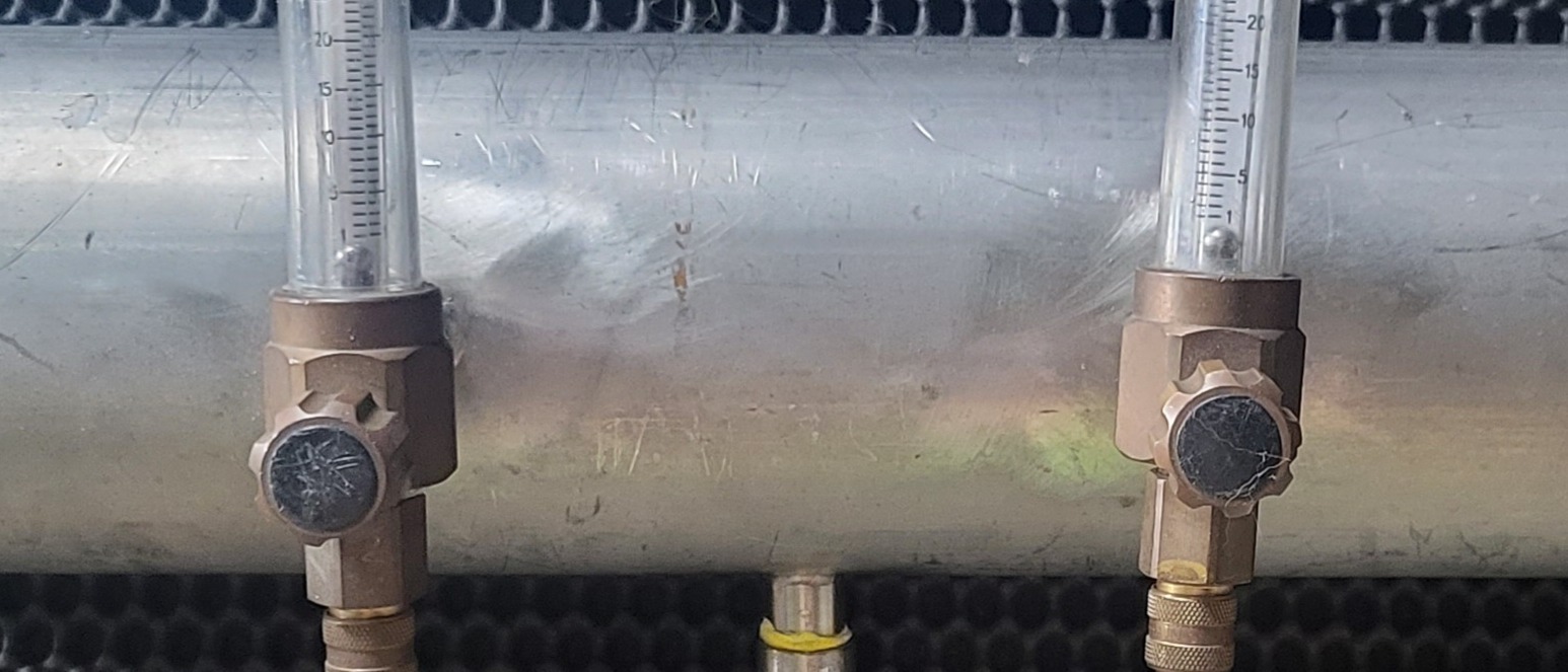 Zbliżenie na dwa rotametry z metalowymi korpusami i pokrętłami regulacyjnymi, zamontowane na metalowej rurze, z widocznymi skalami pomiarowymi i kulkami wskazującymi przepływ.