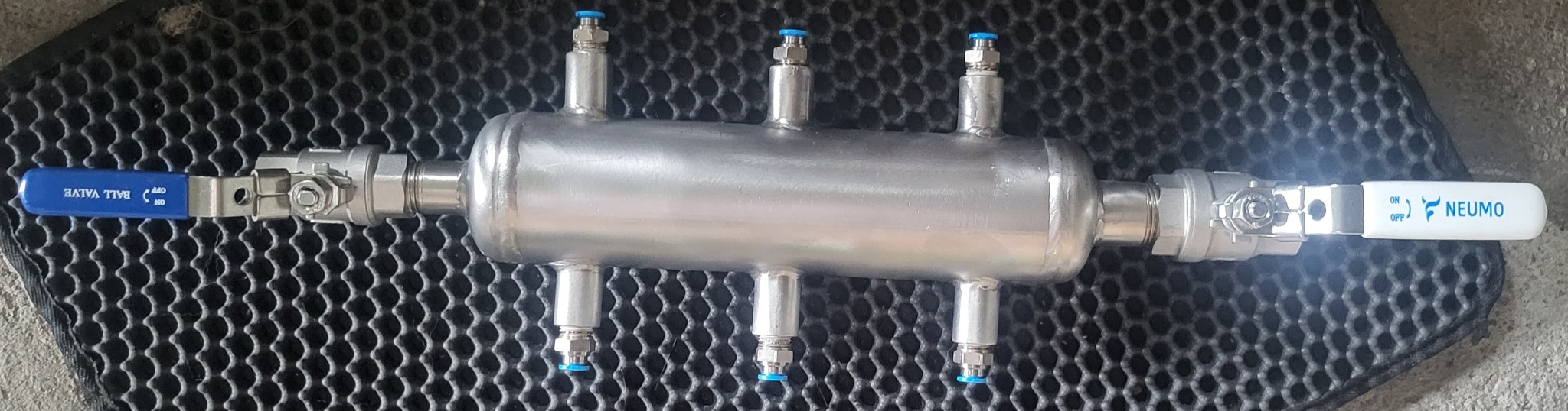 Zbiornik wyrównawczy ze stali nierdzewnej z czterema króćcami i zaworami kulowymi po bokach, leżący na czarnej, gumowej macie.