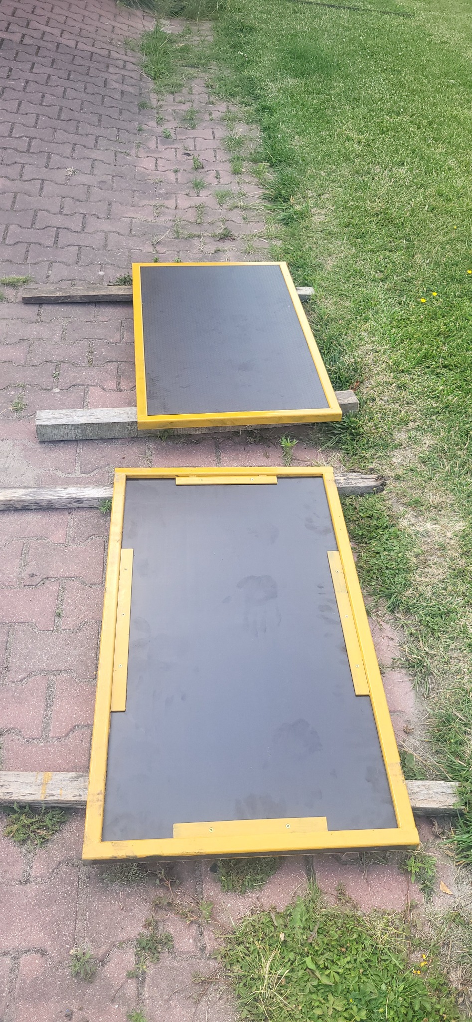 Dwa czarne panele z żółtą metalową ramą, leżące na drewnianych belkach na tle kostki brukowej i trawy.