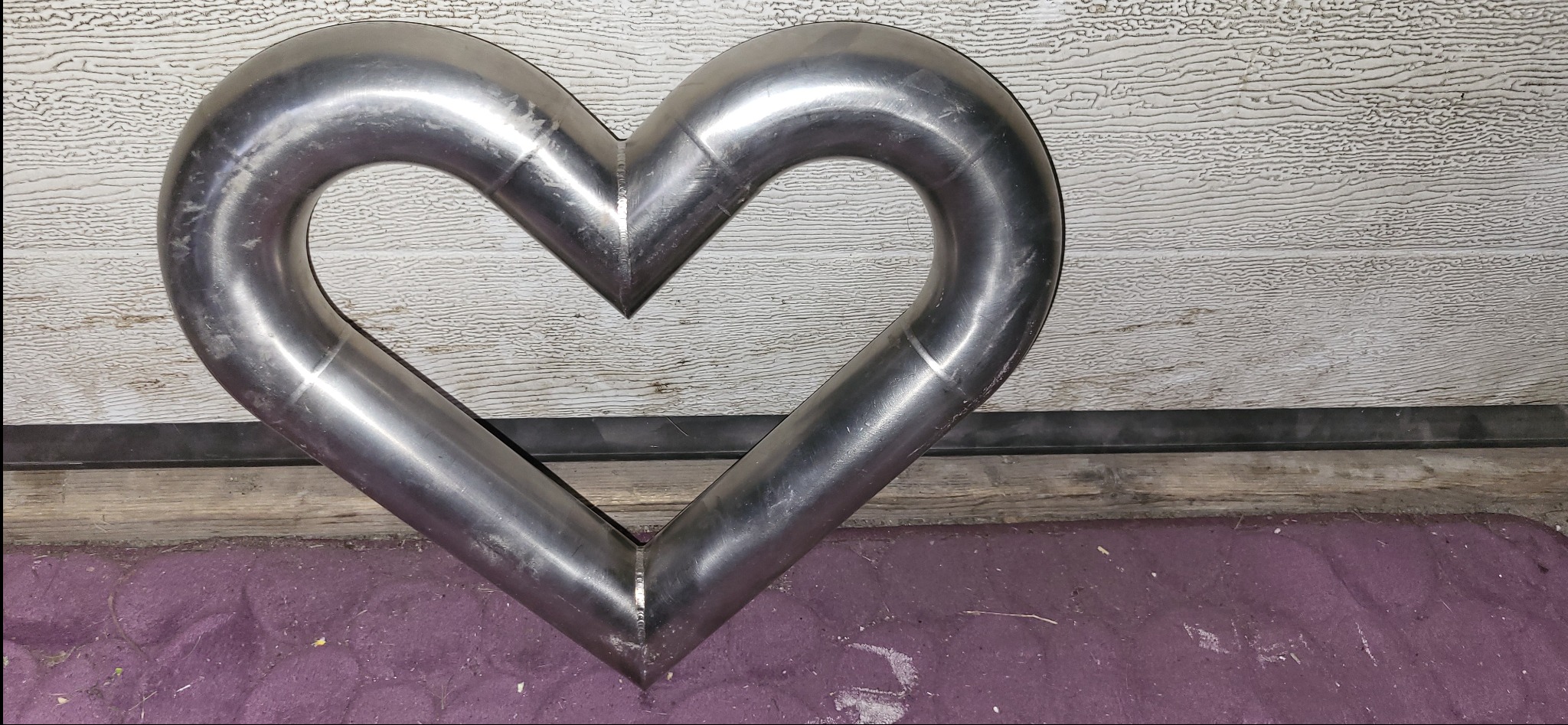 Serce uformowane z giętej, spawanej rury ze stali nierdzewnej, leżące na fioletowej powierzchni, z widocznymi śladami spawania i obróbki metalu.