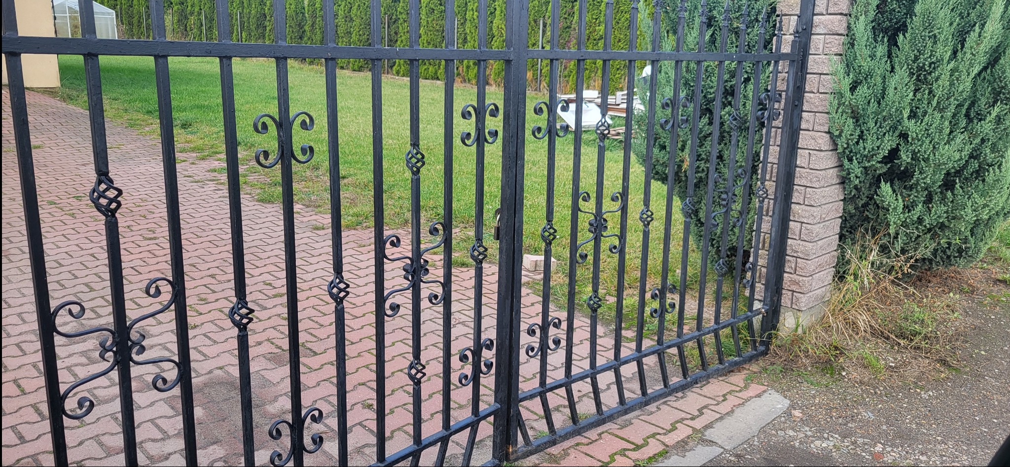 Czarna metalowa brama z ozdobnymi elementami w kształcie spirali i wolut, przylegająca do ceglanego słupka i zielonego żywopłotu, z widoczną kostką brukową i trawnikiem w tle.