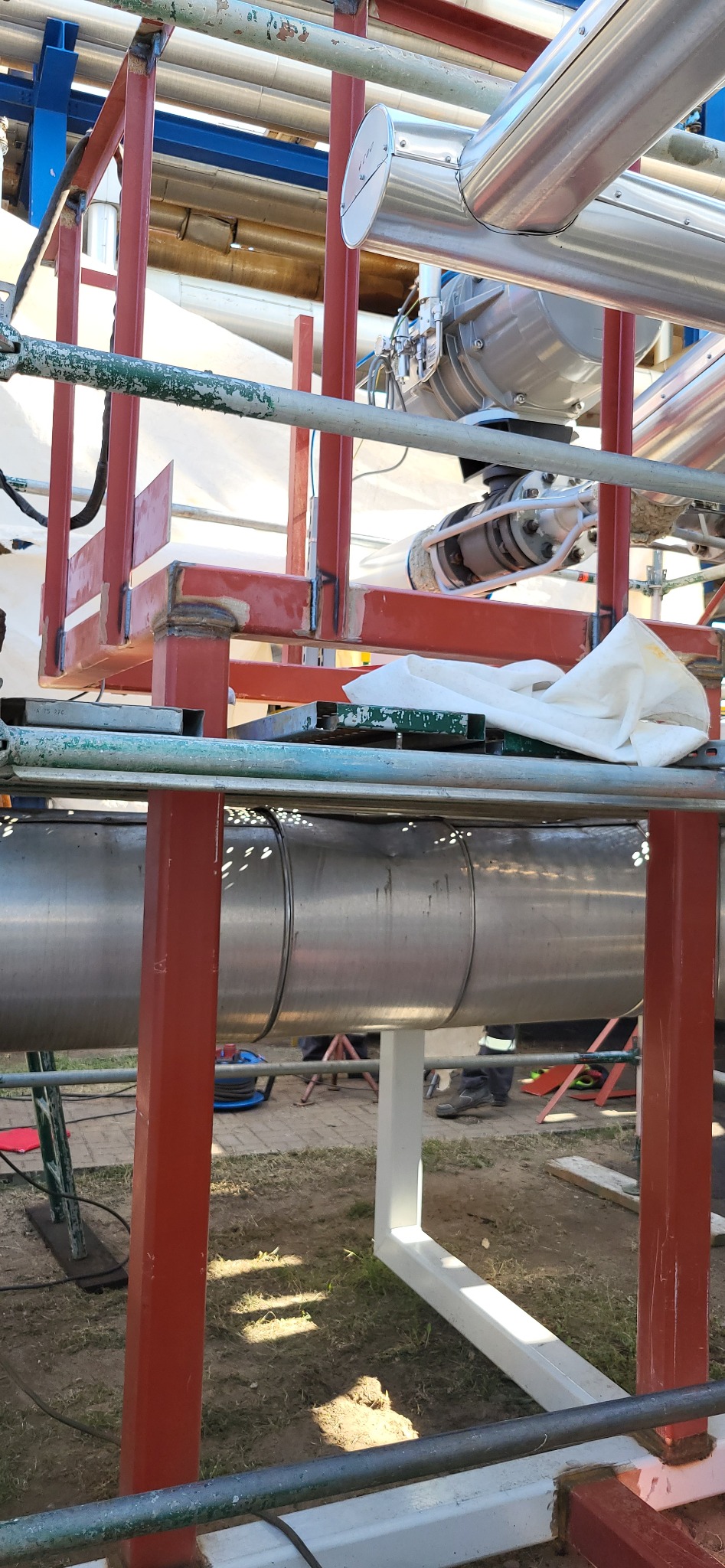 Konstrukcja z metalowych rur i profili, częściowo pomalowana na czerwono i biało, z widocznymi spawami, otoczona rusztowaniem i elementami instalacji przemysłowej.