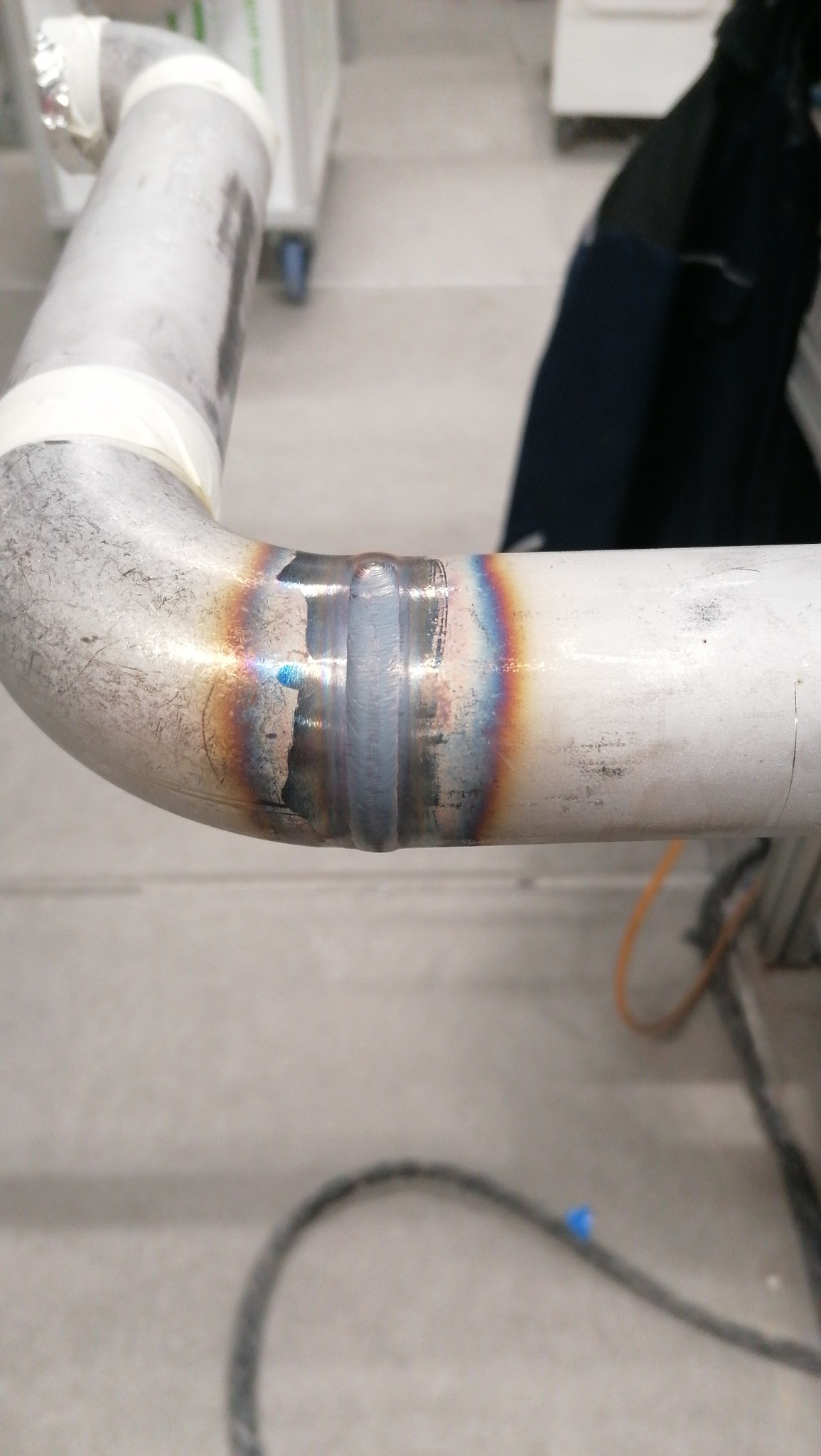 Spoina na zagiętej rurze ze stali nierdzewnej, widoczne tęczowe przebarwienia termiczne, zabezpieczona taśmą.