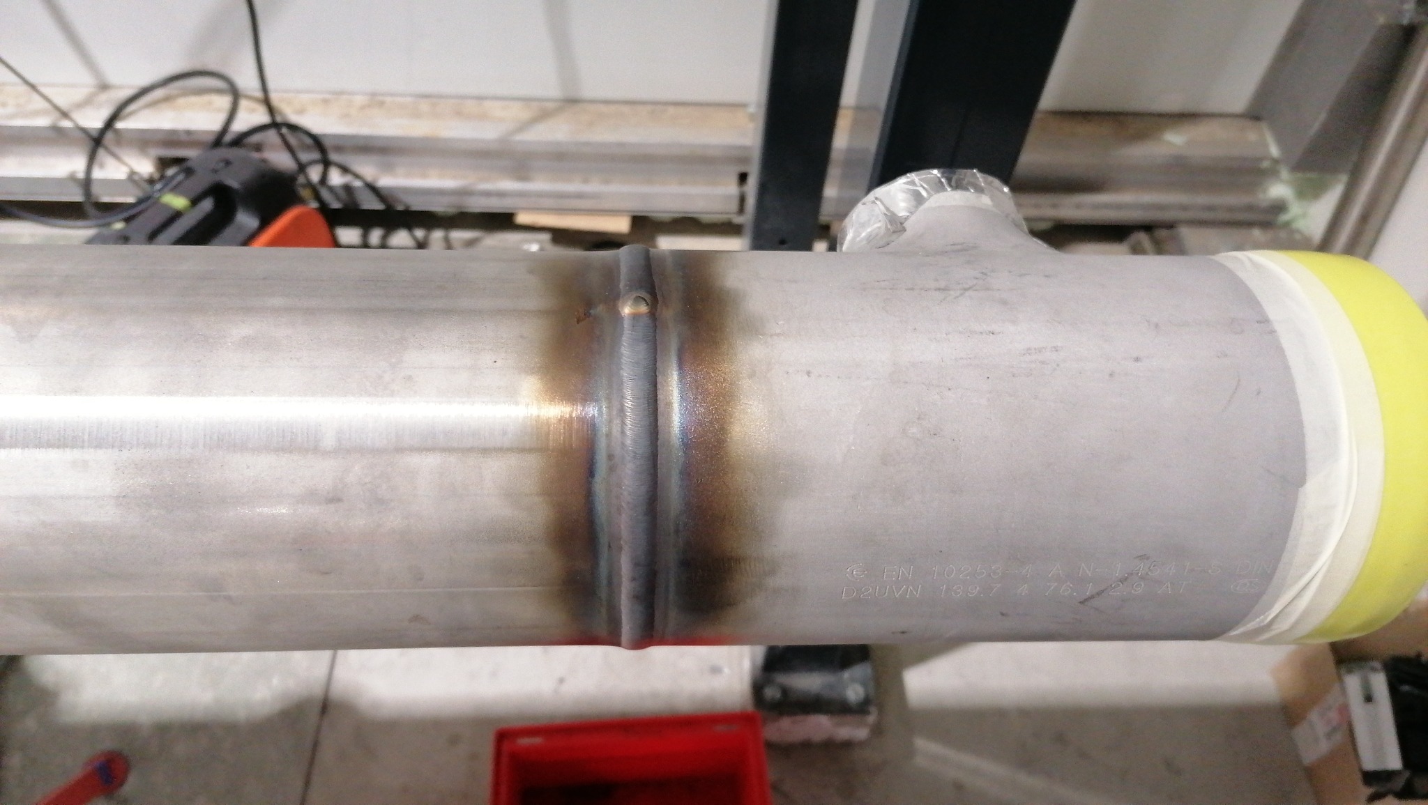 Spoina na rurze ze stali nierdzewnej z widocznymi śladami termicznymi, zabezpieczona taśmą na końcach, zarys narzędzi w tle.