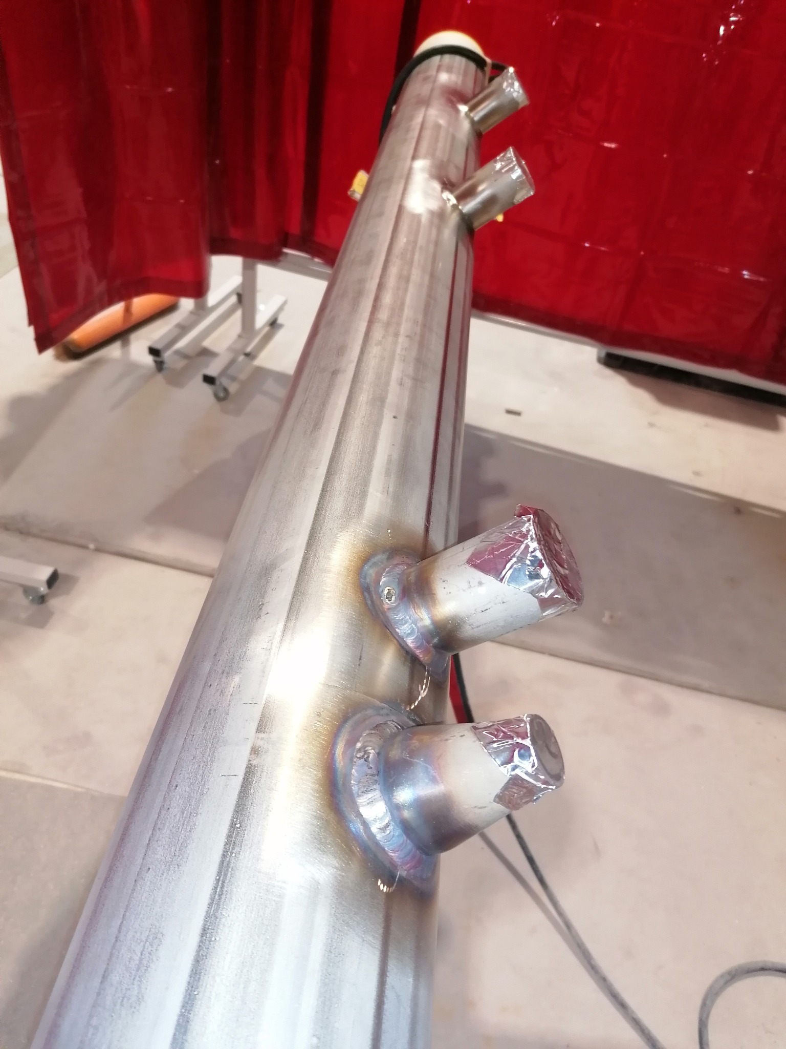 Duży, pionowy zbiornik ze stali nierdzewnej z przyspawanymi króćcami, zabezpieczonymi folią, na tle czerwonej kurtyny spawalniczej i metalowych stojaków.