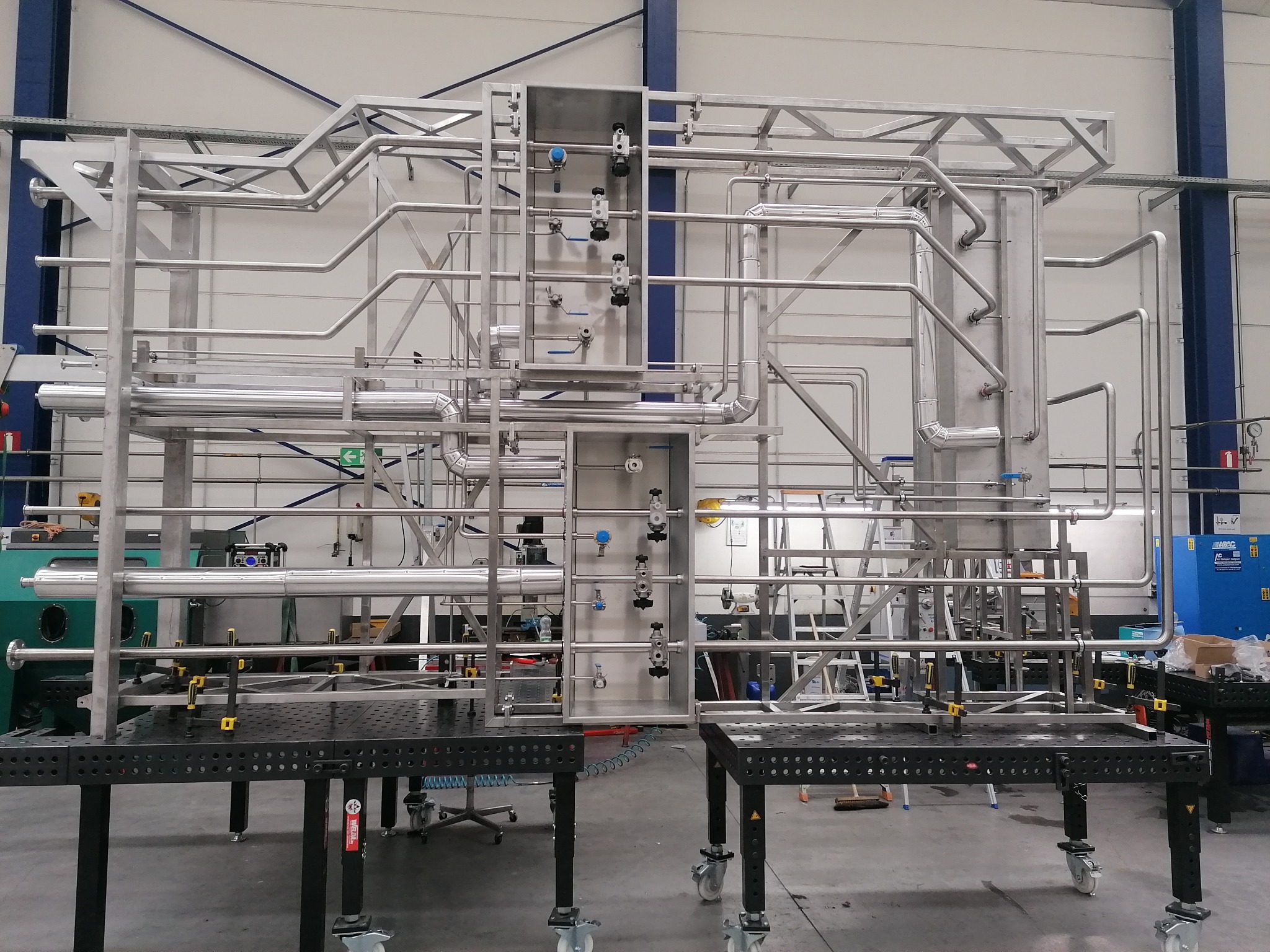 Konstrukcja ze stali nierdzewnej z rurami, zaworami i elementami mocującymi, umieszczona na dwóch stołach spawalniczych z otworami, w hali produkcyjnej.