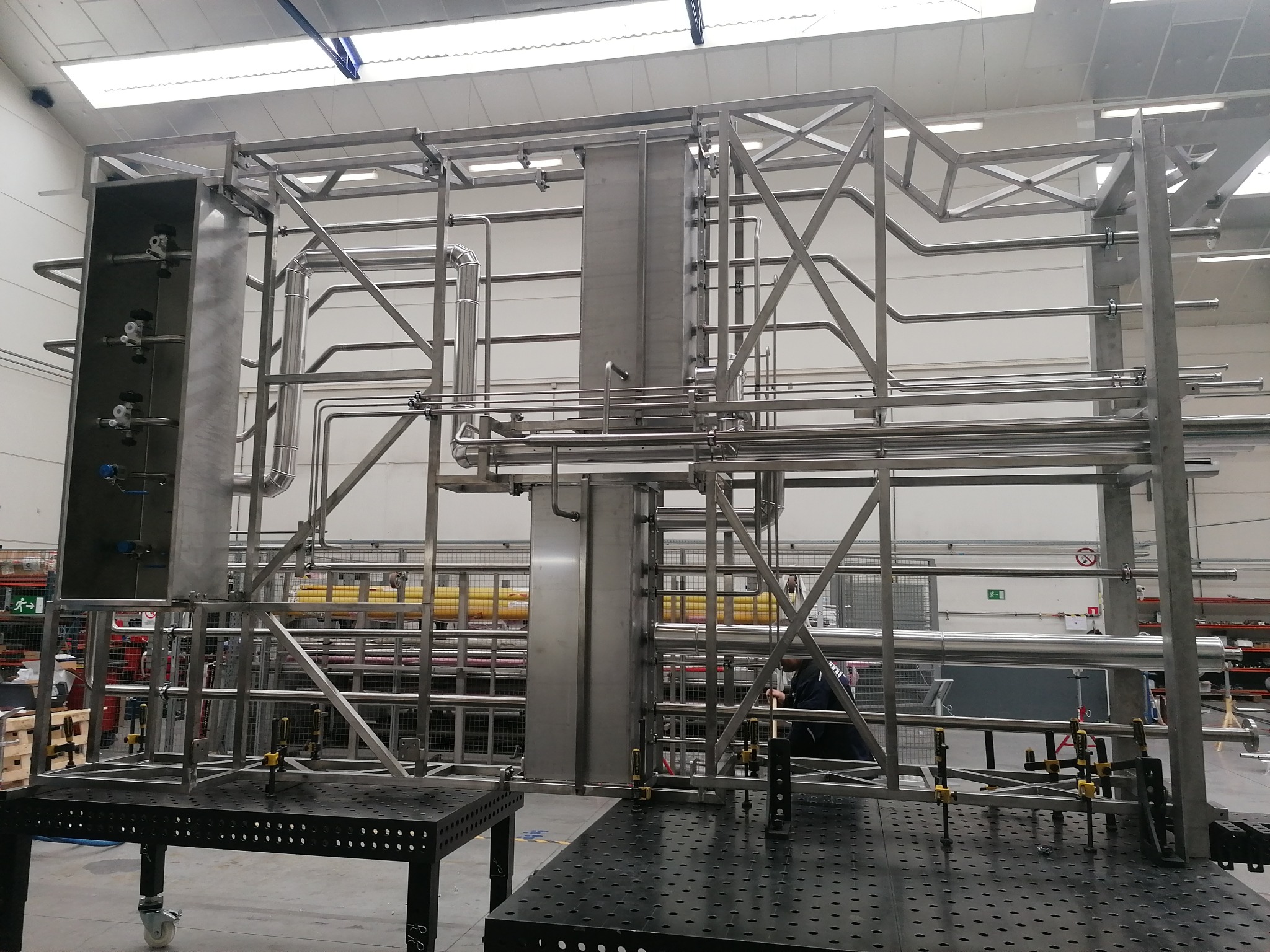 Konstrukcja ze stali nierdzewnej w hali produkcyjnej, widoczne rury, spawane elementy, system zaworów, stoły spawalnicze z otworami, w tle regały magazynowe i pracownik.