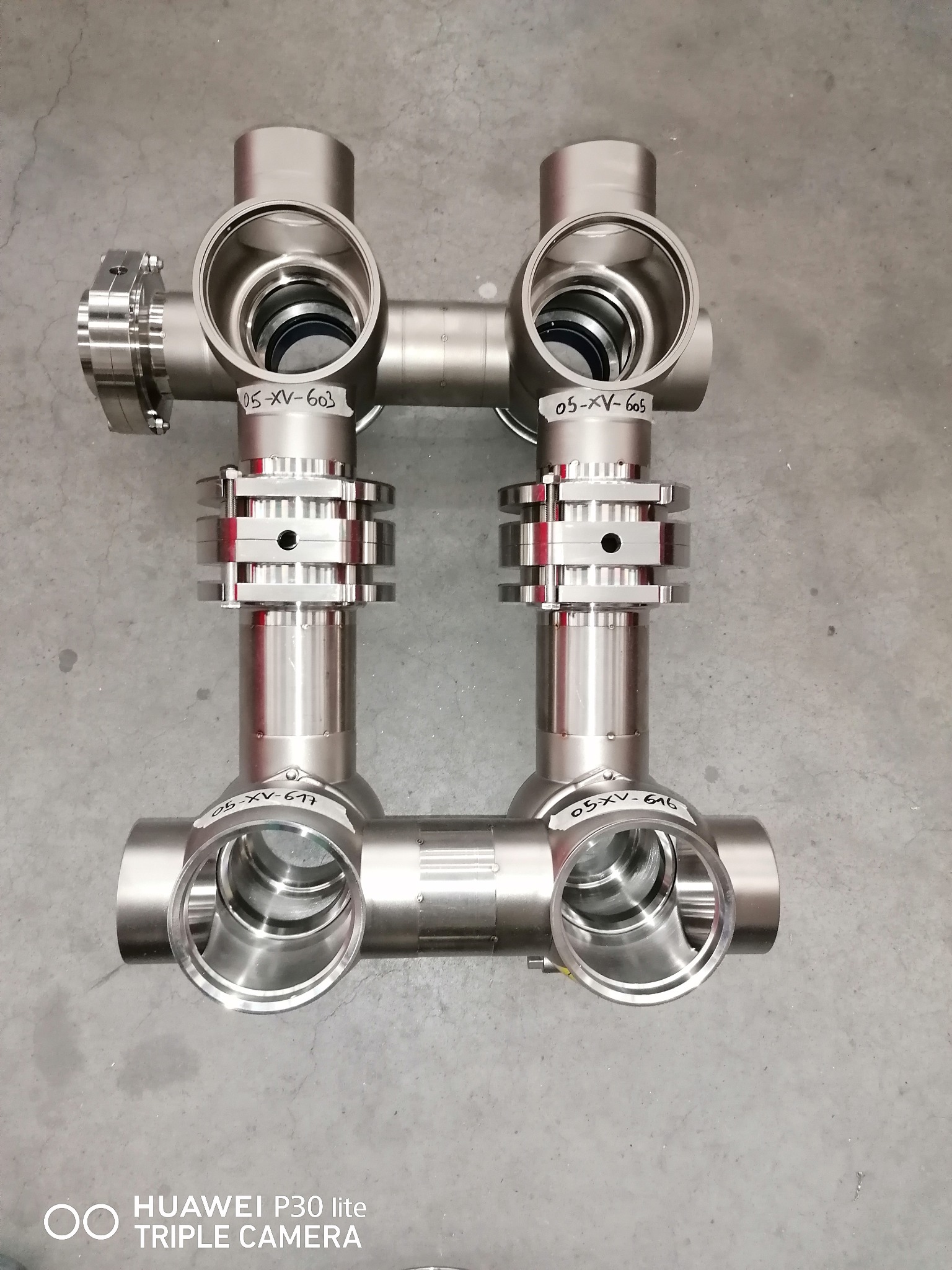 Precyzyjnie wykonane, symetryczne złącze rurowe ze stali nierdzewnej z widocznymi oznaczeniami na elementach, leżące na szarym, betonowym podłożu.