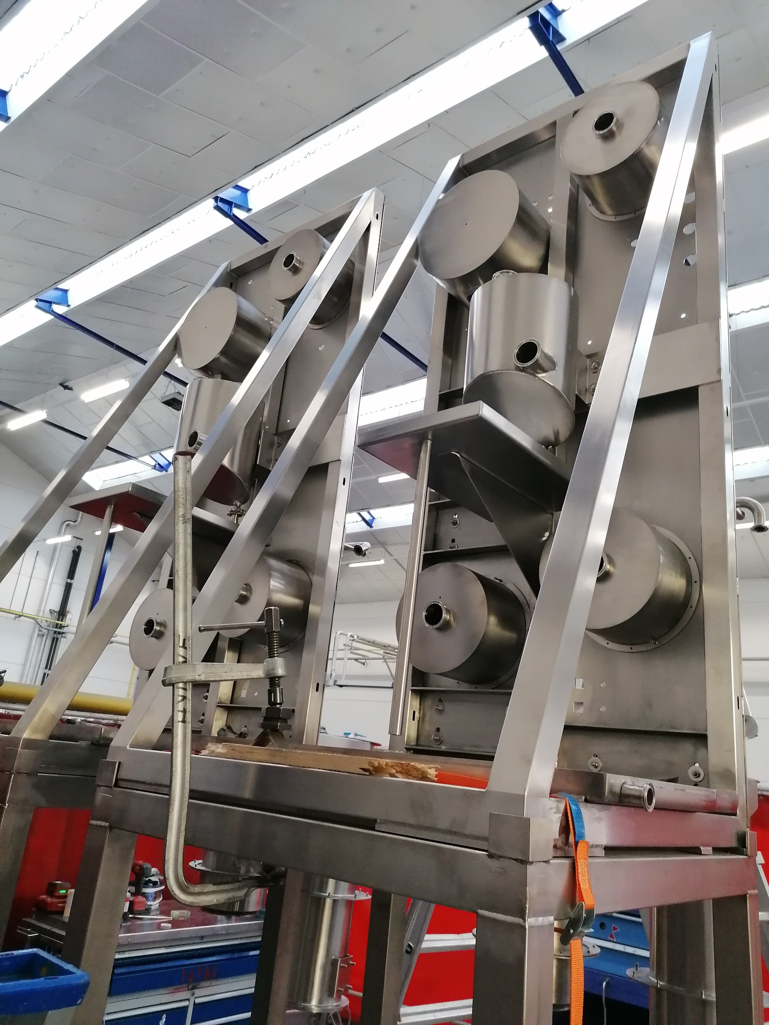 Konstrukcja ze stali nierdzewnej z cylindrycznymi elementami, widoczna w hali produkcyjnej, częściowo zmontowana, z zaciskiem śrubowym i pasem transportowym, oświetlona światłem dziennym.