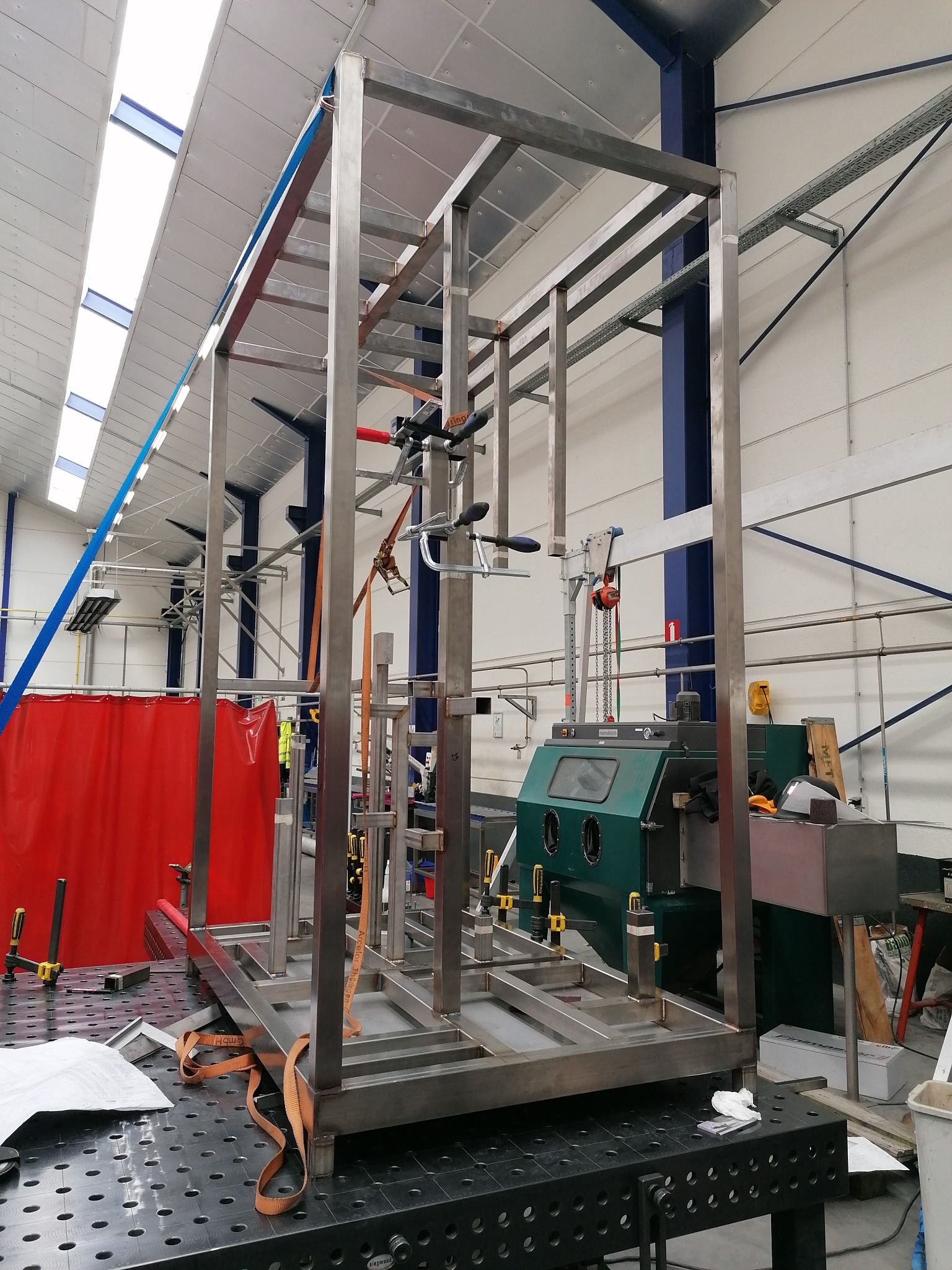 Stalowa rama konstrukcyjna w trakcie budowy w warsztacie, zabezpieczona pasami transportowymi, widoczne narzędzia i wyposażenie warsztatowe w tle.