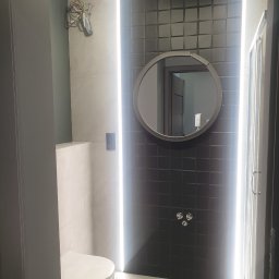 Letex - Nowoczesna łazienka z czarną, teksturowaną ścianą, okrągłym lustrem z podświetleniem LED, wiszącą toaletą i kabiną prysznicową; widoczne wystające przewody elektryczne.