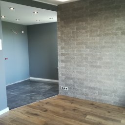 Letex - Wykończone wnętrze mieszkania z szarą, dekoracyjną ścianą imitującą kamień, drewnianą podłogą i nowoczesną lampą sufitową w kształcie sześcianu, widoczne gniazdka elektryczne.