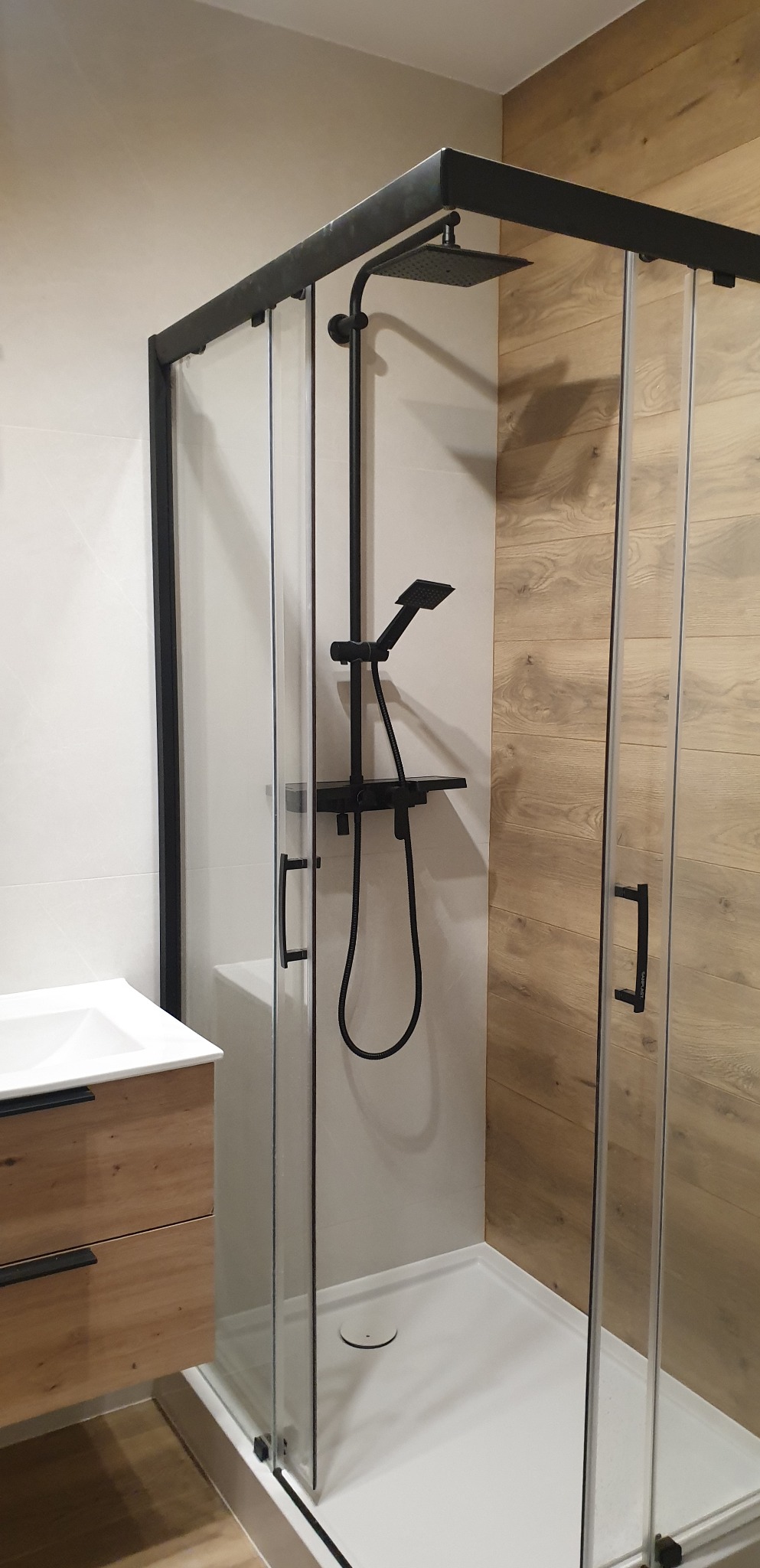 Nowoczesna kabina prysznicowa z czarnymi profilami i deszczownicą w łazience z drewnopodobnymi płytkami, widoczna umywalka z szafką o froncie w kolorze jasnego drewna.