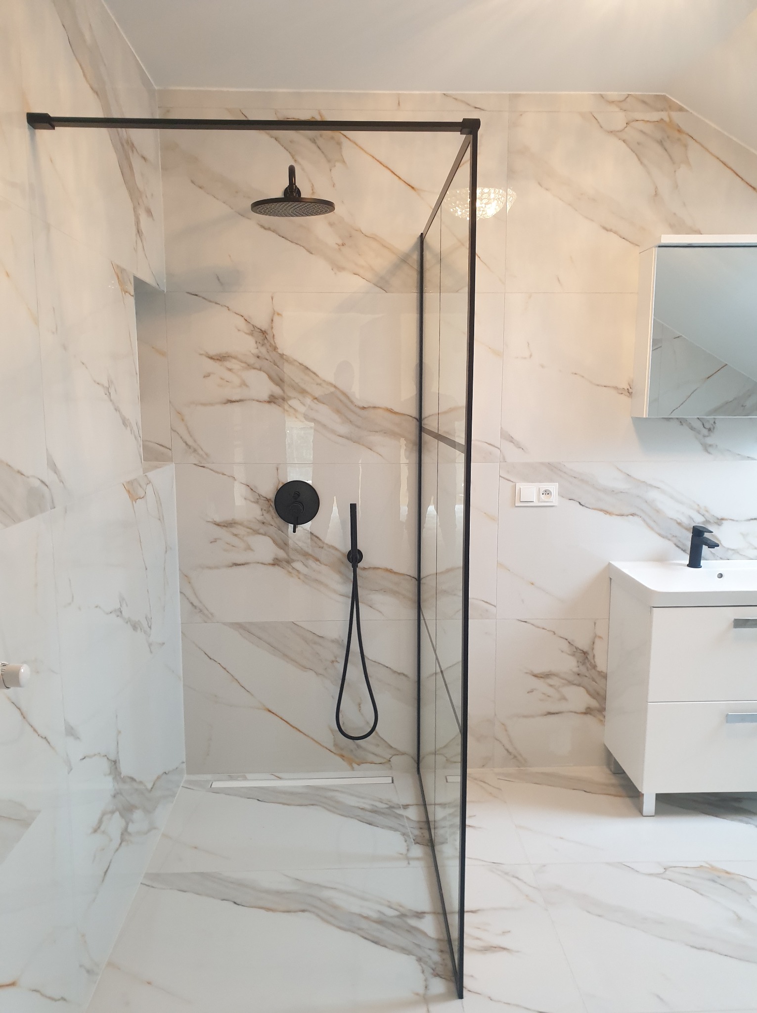 Nowoczesna łazienka z kabiną prysznicową typu walk-in, wykończona białymi płytkami imitującymi marmur z delikatnymi złotymi żyłami, czarna armatura, biała szafka z umywalką podwieszana...