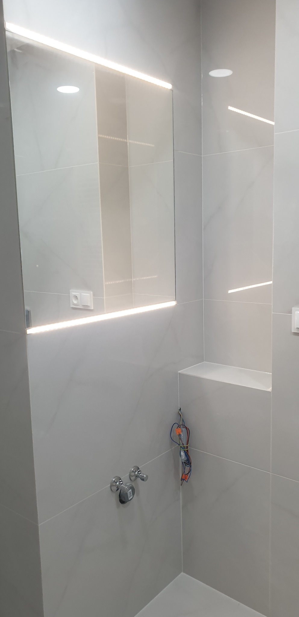 Nowoczesna łazienka z szarymi płytkami, podświetlanym lustrem i wnęką, widoczne przewody elektryczne przed podłączeniem.