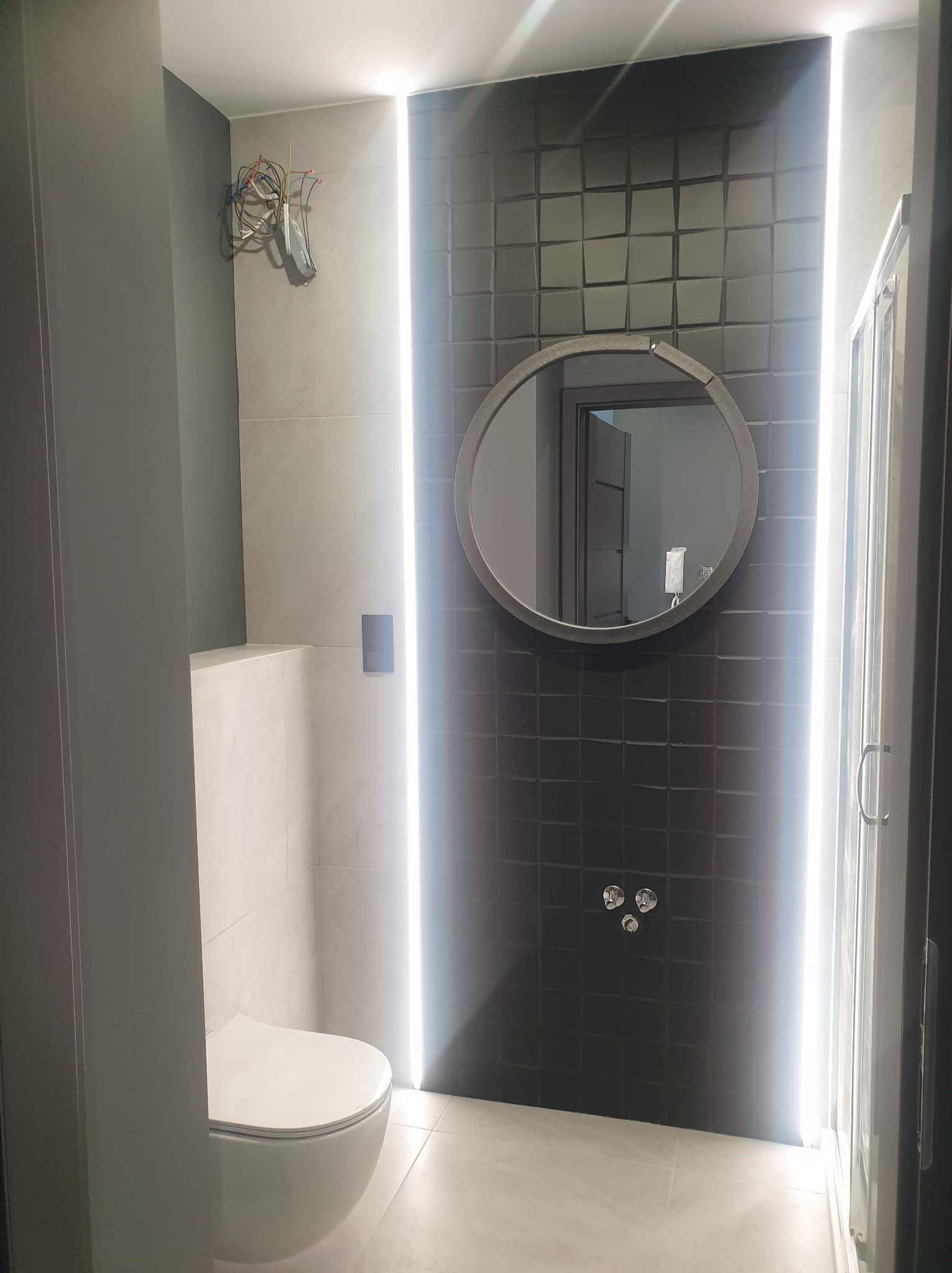 Nowoczesna łazienka z czarną, teksturowaną ścianą, okrągłym lustrem z podświetleniem LED, wiszącą toaletą i kabiną prysznicową; widoczne wystające przewody elektryczne.