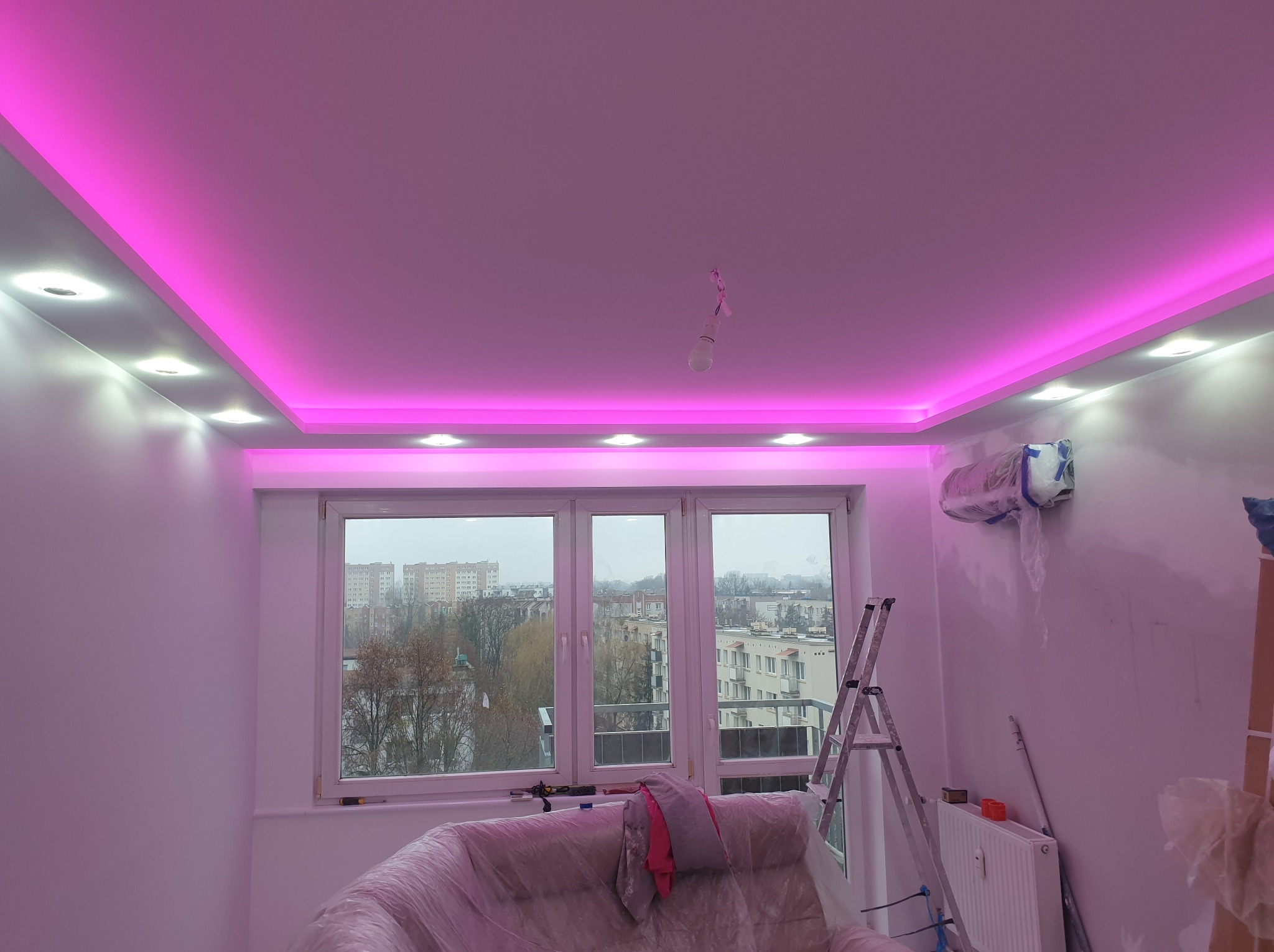 Pokój w trakcie remontu z podwieszanym sufitem z oświetleniem LED w kolorze różowym, widoczne zabezpieczone meble folią malarską, drabina i narzędzia malarskie.