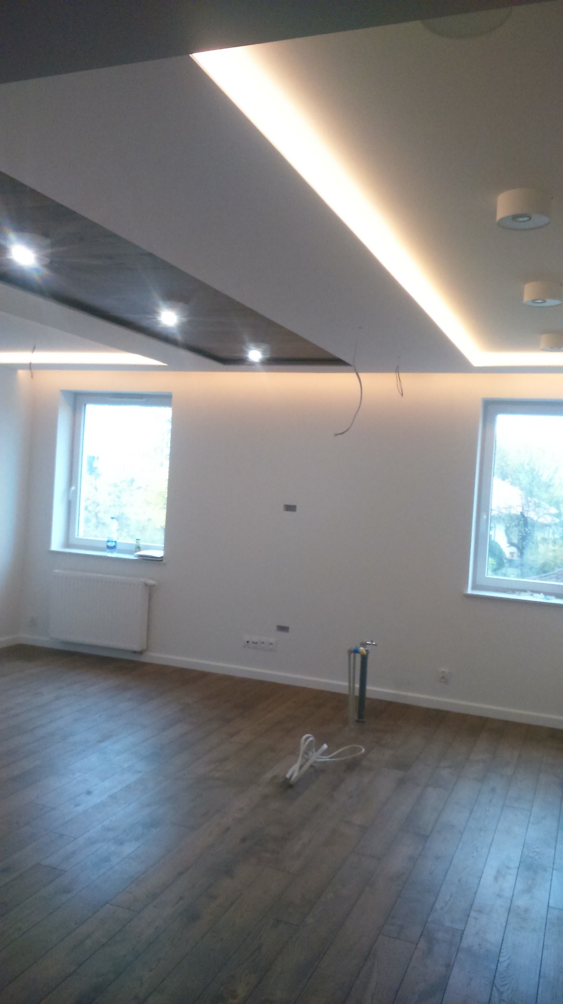 Pokój po remoncie: drewnopodobna podłoga, białe ściany, sufit z oświetleniem LED i elementami dekoracyjnymi, widoczne okna z grzejnikiem i wystającymi kablami.