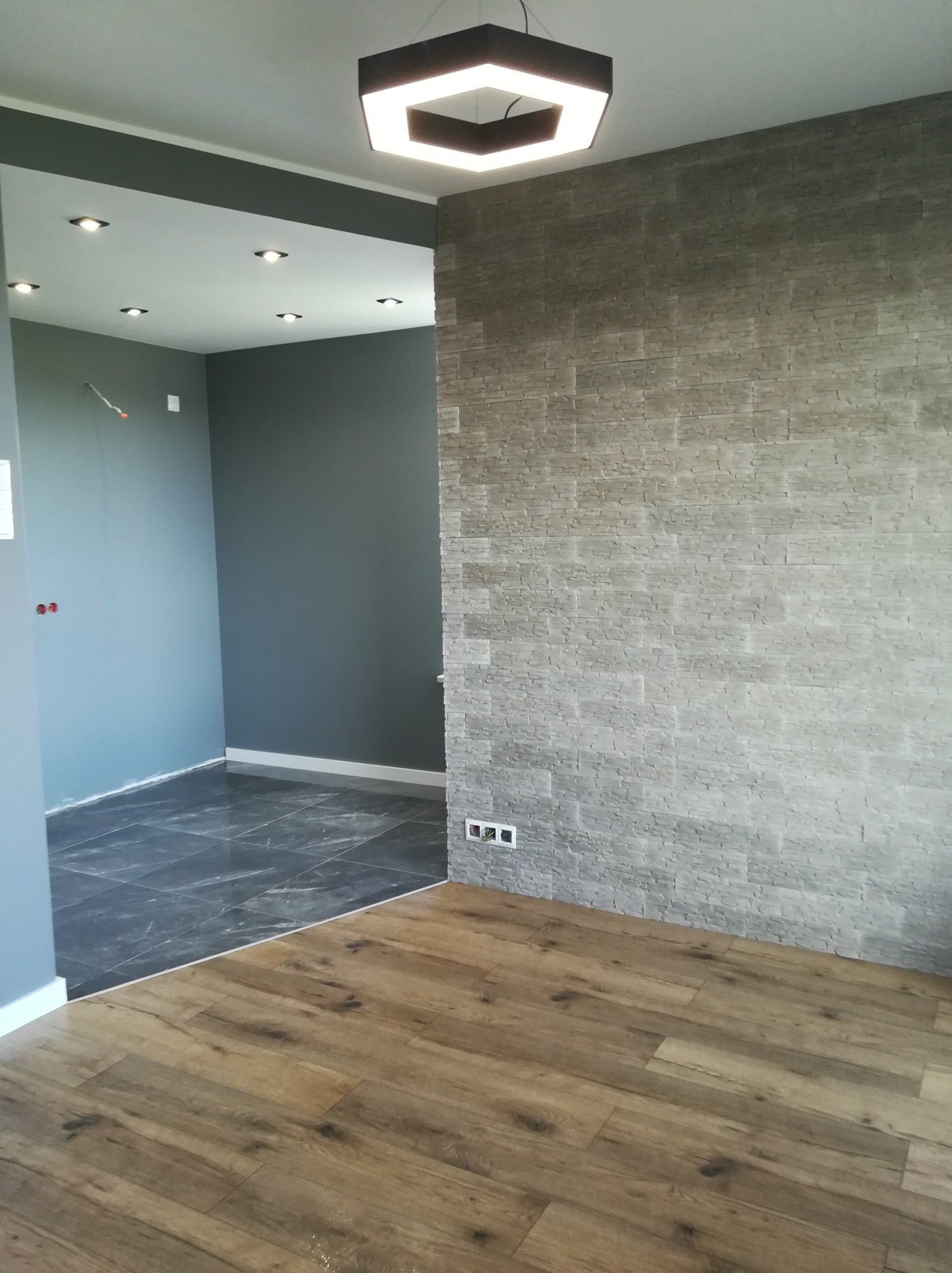 Wykończone wnętrze mieszkania z szarą, dekoracyjną ścianą imitującą kamień, drewnianą podłogą i nowoczesną lampą sufitową w kształcie sześcianu, widoczne gniazdka elektryczne.