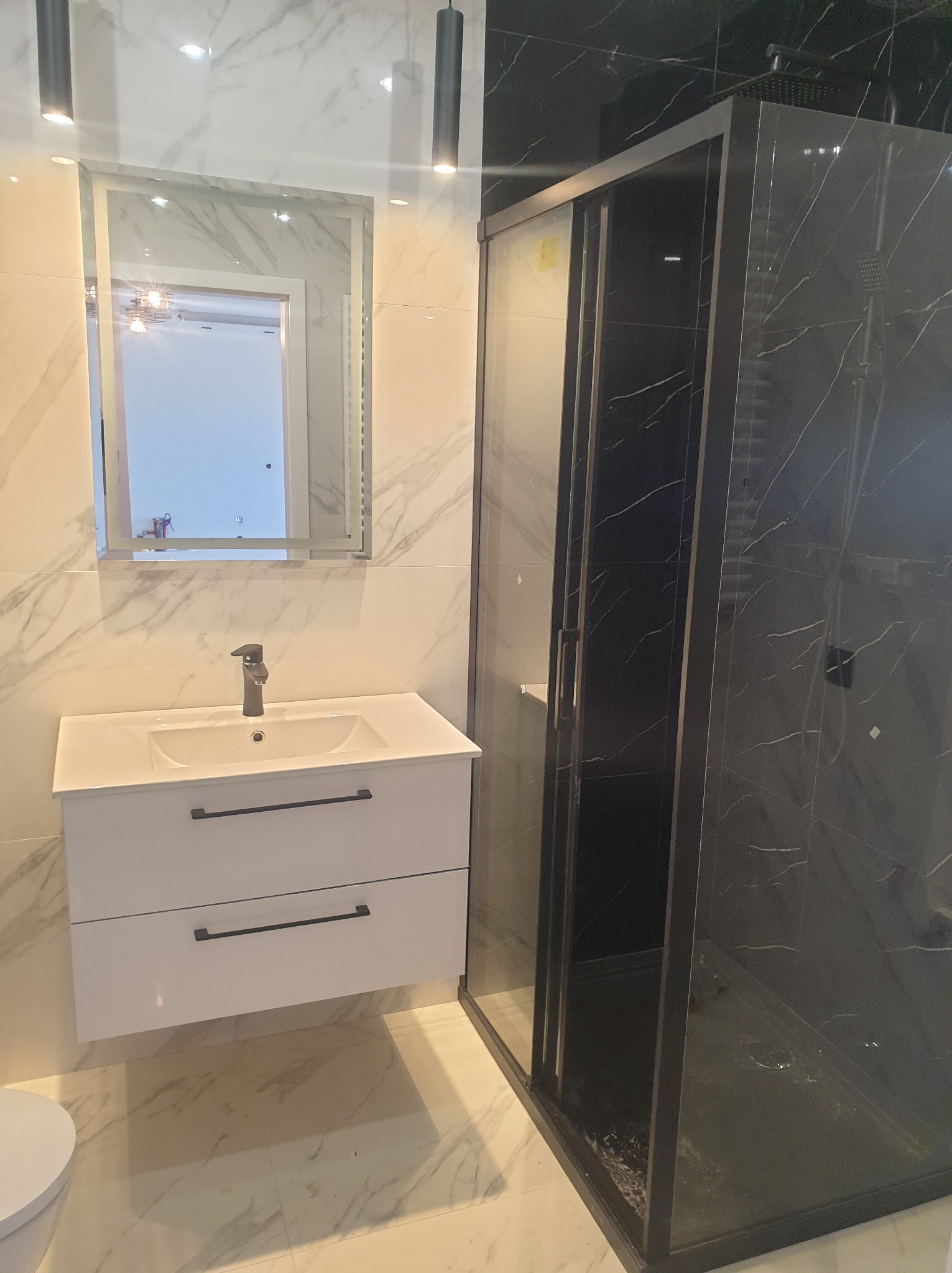 Nowoczesna łazienka z biało-czarnymi marmurowymi płytkami, białą szafką pod umywalką z czarnymi uchwytami, lustrem z podświetleniem LED i czarną kabiną prysznicową z deszczownicą.