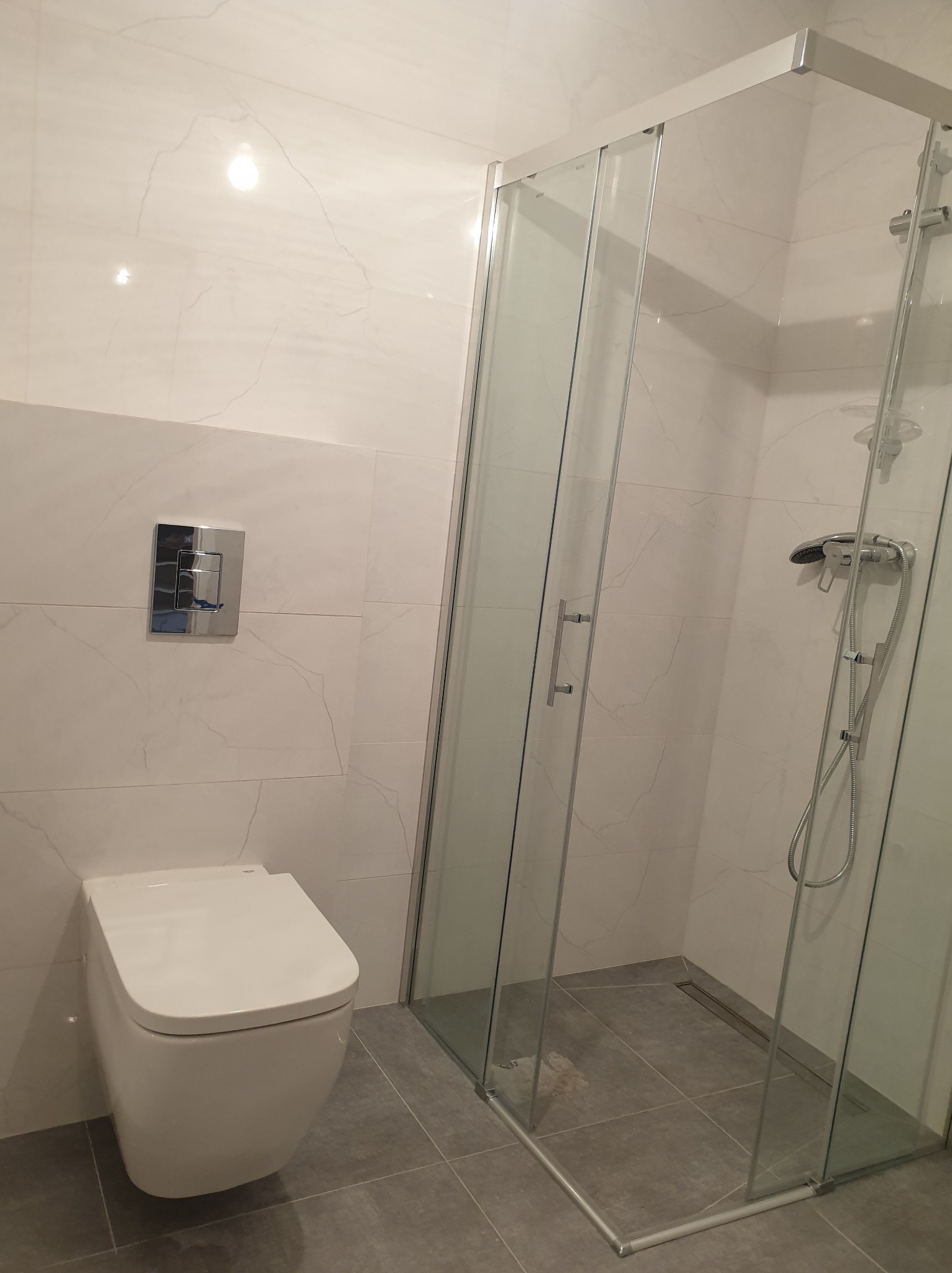 Nowoczesna łazienka z białymi marmurkowymi płytkami, podwieszaną toaletą i kabiną prysznicową ze szklanymi drzwiami, widok z boku.