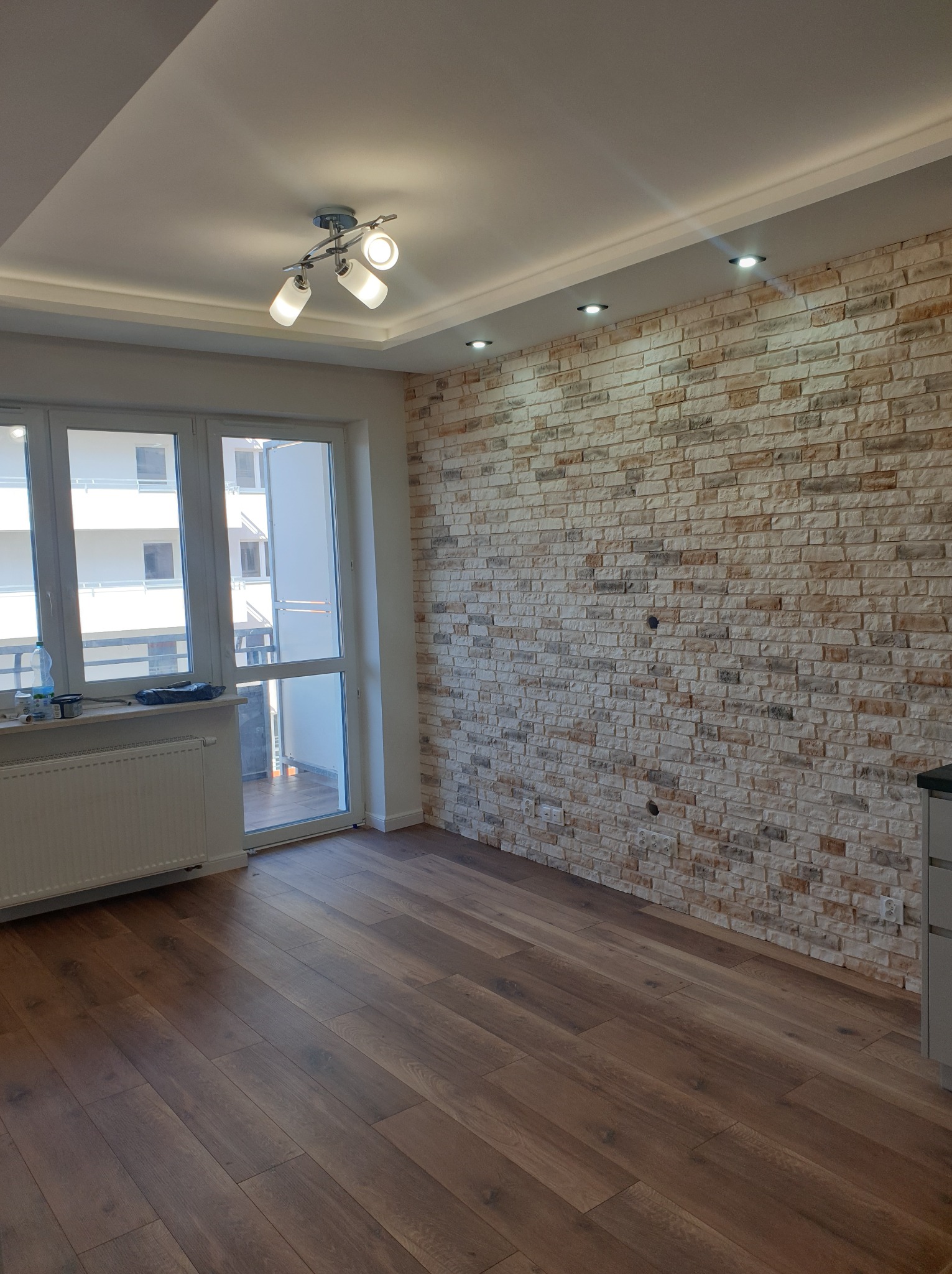 Wykończone wnętrze w Białymstoku: ściana z dekoracyjnej cegły, drewniana podłoga, okno balkonowe z widokiem na budynek, nowoczesne oświetlenie sufitowe.