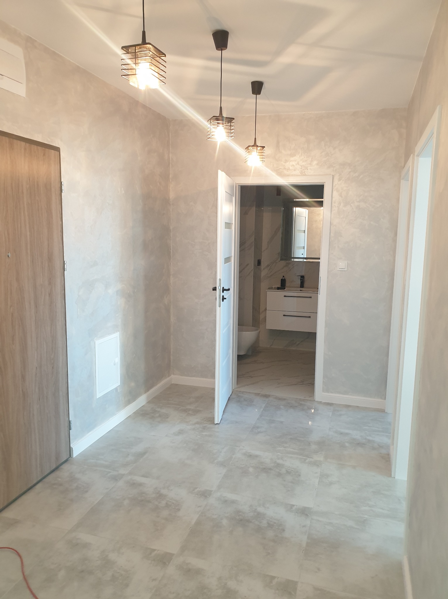 Wykończony korytarz z otwartymi drzwiami do łazienki, widoczne szare płytki podłogowe, ściany z efektem dekoracyjnym, trzy wiszące lampy z metalowymi kloszami i fragment instalacji elektrycznej...