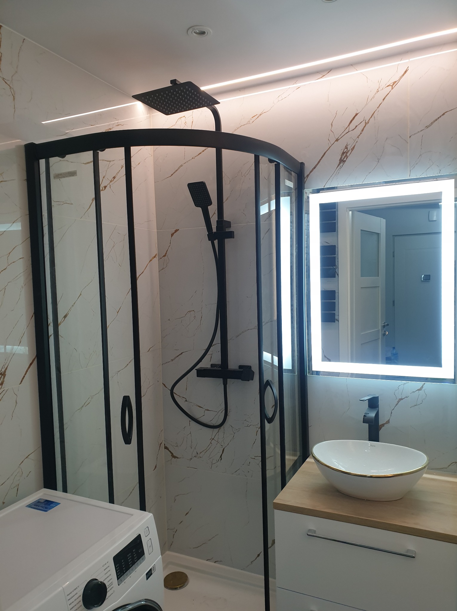 Nowoczesna łazienka z kabiną prysznicową z czarnymi profilami, umywalką nablatową z złotym rantem i podświetlanym lustrem, wykończona płytkami imitującymi marmur z złotymi żyłami.