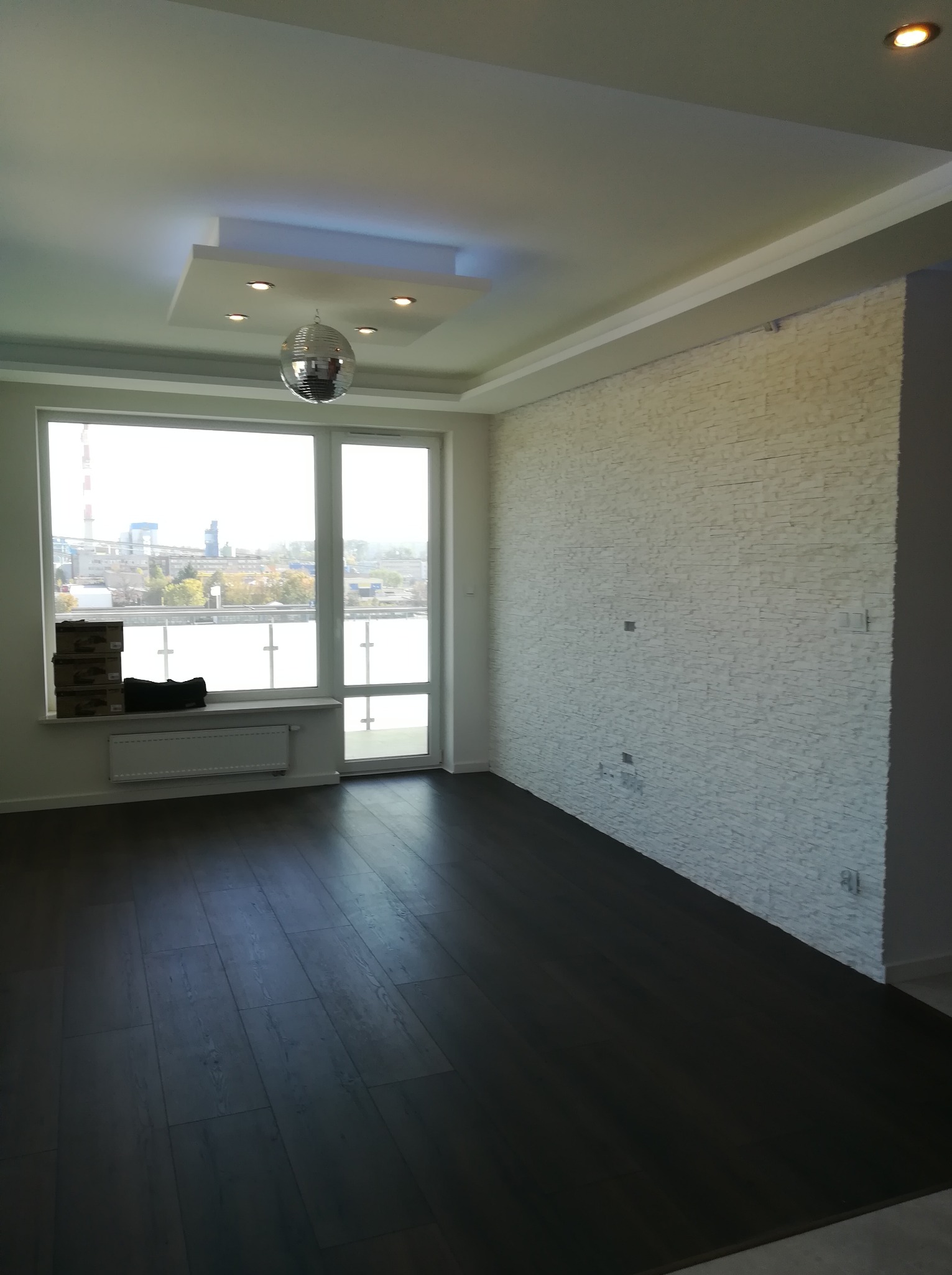 Jasne, przestronne wnętrze po remoncie: ciemna podłoga, ściana wyłożona dekoracyjnym kamieniem, okno z widokiem na zabudowania przemysłowe, srebrna kula dyskotekowa zwisająca z sufitu...