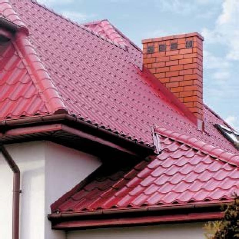 Czerwony dach z blachodachówki w kształcie fali, z widocznym kominem z cegły i rynnami w kolorze dachu, na tle błękitnego nieba.