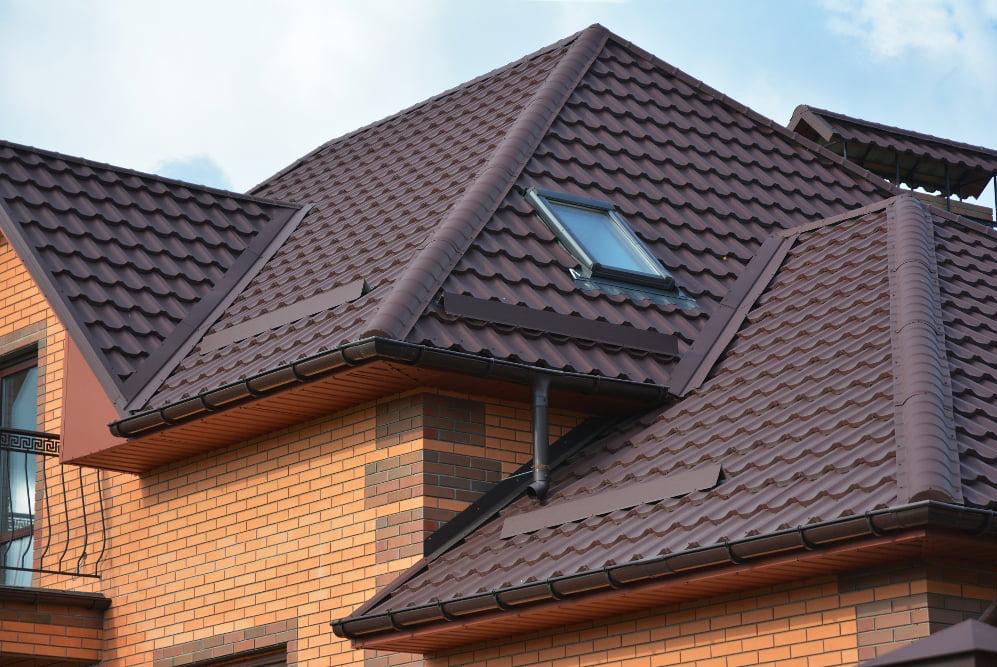 Brązowy dach z blachodachówki z oknem dachowym i rynnami, widok na fragment elewacji z cegły klinkierowej.