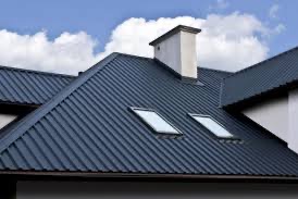 Nowoczesny dach z blachy trapezowej w kolorze grafitowym, z dwoma oknami dachowymi i murowanym kominem na tle błękitnego nieba z białymi chmurami.