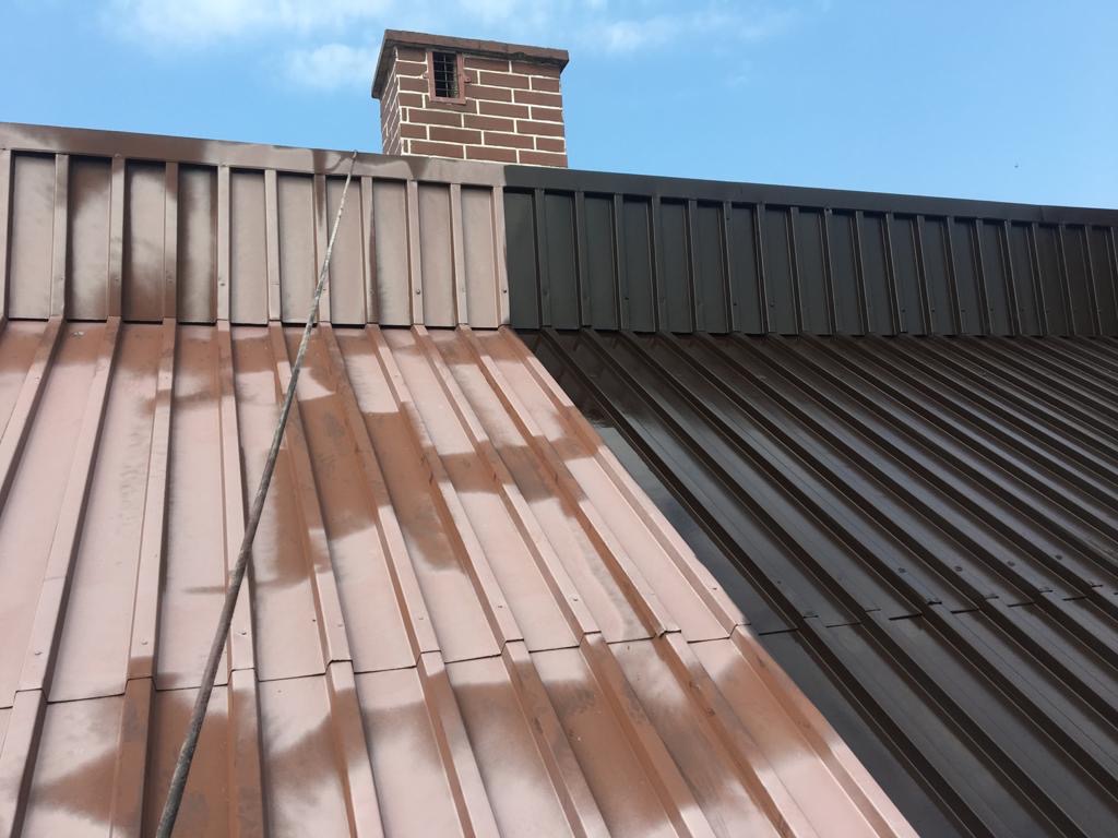 Częściowo pomalowany blaszany dach w kolorze brązowym, z widocznym kominem z cegły w tle i liną zwisającą z góry.