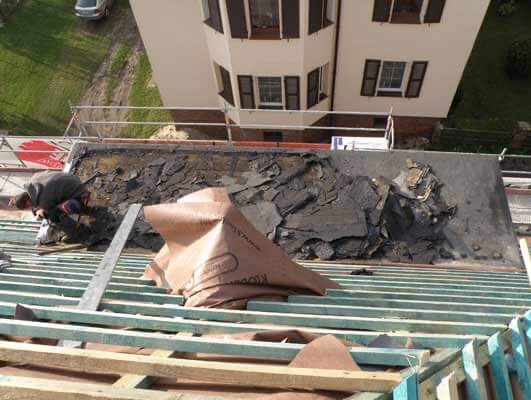 Demontaż starego pokrycia dachowego z papy na drewnianej konstrukcji więźby, widoczny pracownik w trakcie pracy oraz fragment elewacji budynku.