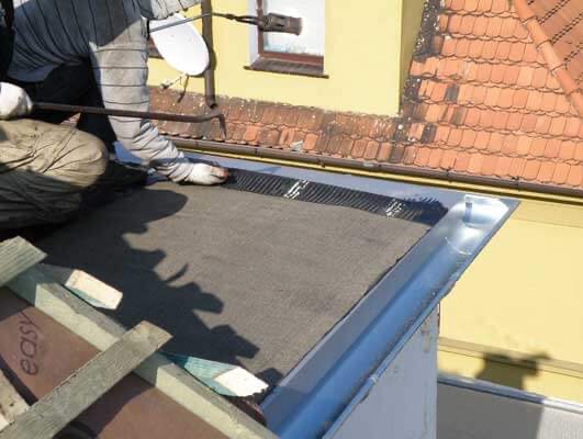 Pracownik w białych rękawicach montuje papę bitumiczną na dachu, widoczna nowa metalowa rynna i fragment dachu sąsiedniego budynku z pomarańczową dachówką.