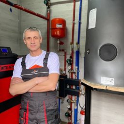 MAZUR SERVICE SPÓŁKA Z OGRANICZONĄ ODPOWIEDZIALNOŚCIĄ - Instalator w kombinezonie roboczym z założonymi rękami na tle nowej instalacji grzewczej z zbiornikiem akumulacyjnym i czerwonym naczyniem wzbiorczym.