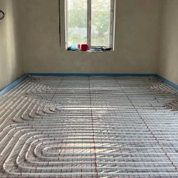 MAZUR SERVICE SPÓŁKA Z OGRANICZONĄ ODPOWIEDZIALNOŚCIĄ - Białe rurki ogrzewania podłogowego ułożone na czerwonej siatce w surowym pomieszczeniu z oknem i metalowym rusztem pod sufitem.