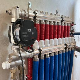 MAZUR SERVICE SPÓŁKA Z OGRANICZONĄ ODPOWIEDZIALNOŚCIĄ - Rozdzielacz ogrzewania podłogowego z pompą obiegową, rurami w kolorze czerwonym i niebieskim oraz białymi zaworami termostatycznymi, zamontowany na szarej ścianie.