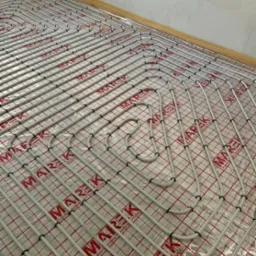 Białe rurki ogrzewania podłogowego ułożone na folii z czerwoną kratką i logotypem producenta, widok z góry, surowy stan ściany w tle.