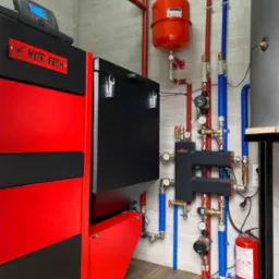 Zainstalowany, nowoczesny piec Metal-Fach z podajnikiem, widoczne rury instalacji grzewczej w kolorach czerwonym i niebieskim oraz zbiornik wyr&oacute;wnawczy i gaśnica.