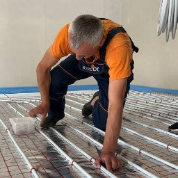 MAZUR SERVICE SPÓŁKA Z OGRANICZONĄ ODPOWIEDZIALNOŚCIĄ - Instalator w kombinezonie roboczym układa białe rurki ogrzewania podłogowego na folii z czerwoną siatką, przymocowując je plastikowymi elementami.