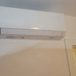 Biały klimatyzator wiszący marki Gree zamontowany na ścianie w jasnym pomieszczeniu, widoczny fragment instalacji odprowadzającej skropliny i narożnik futryny drzwiowej.