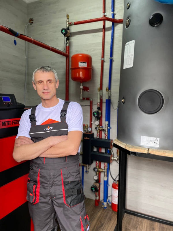 Instalator w kombinezonie roboczym z założonymi rękami na tle nowej instalacji grzewczej z zbiornikiem akumulacyjnym i czerwonym naczyniem wzbiorczym.