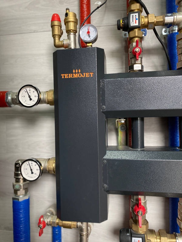 Szczegółowe ujęcie węzła cieplnego Termojet z manometrami, zaworami i rurami, zamontowanego na ścianie z panelami imitującymi drewno.