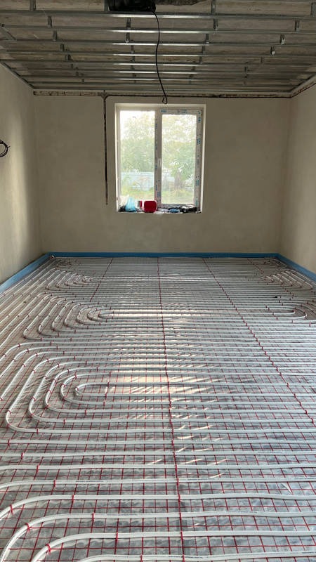 Białe rurki ogrzewania podłogowego ułożone na czerwonej siatce w surowym pomieszczeniu z oknem i metalowym rusztem pod sufitem.