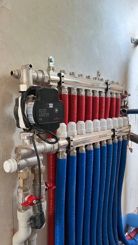 Rozdzielacz ogrzewania podłogowego z pompą obiegową, rurami w kolorze czerwonym i niebieskim oraz białymi zaworami termostatycznymi, zamontowany na szarej ścianie.